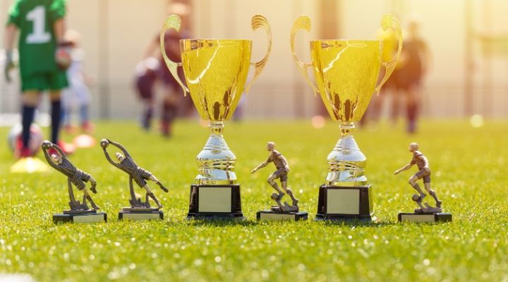 Two gold cup trophies and four smaller trophies laid out on the grass while soccer athletes play on the background