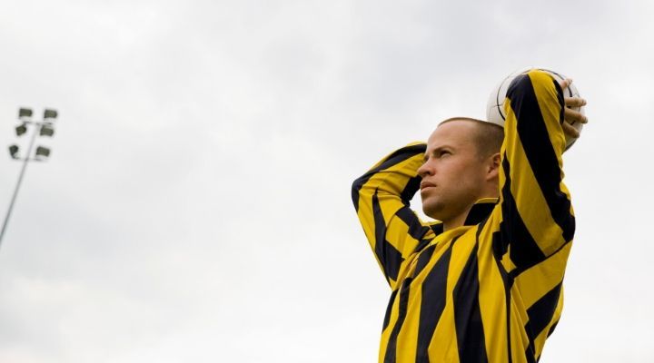 Man wearing yellow and black striped shirt holding the soccer ball above his head and about to throw the ball