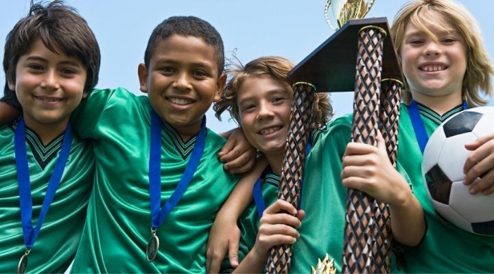 Four young soccer players smiling at the camera while showing off their medals and championship trophy