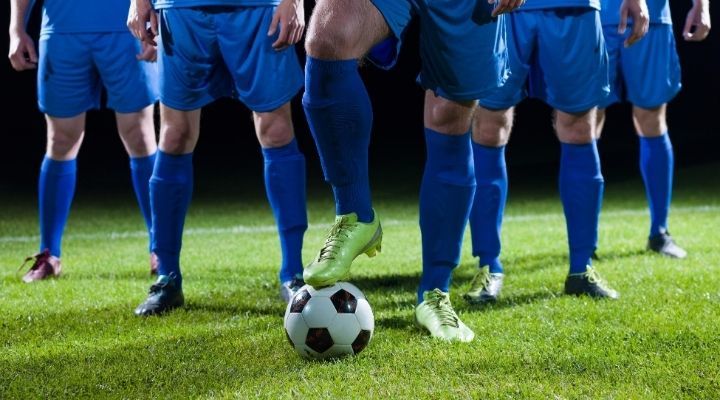 Five soccer players in blue uniform lined up on a field with the center player holding the ball in place with his right foot